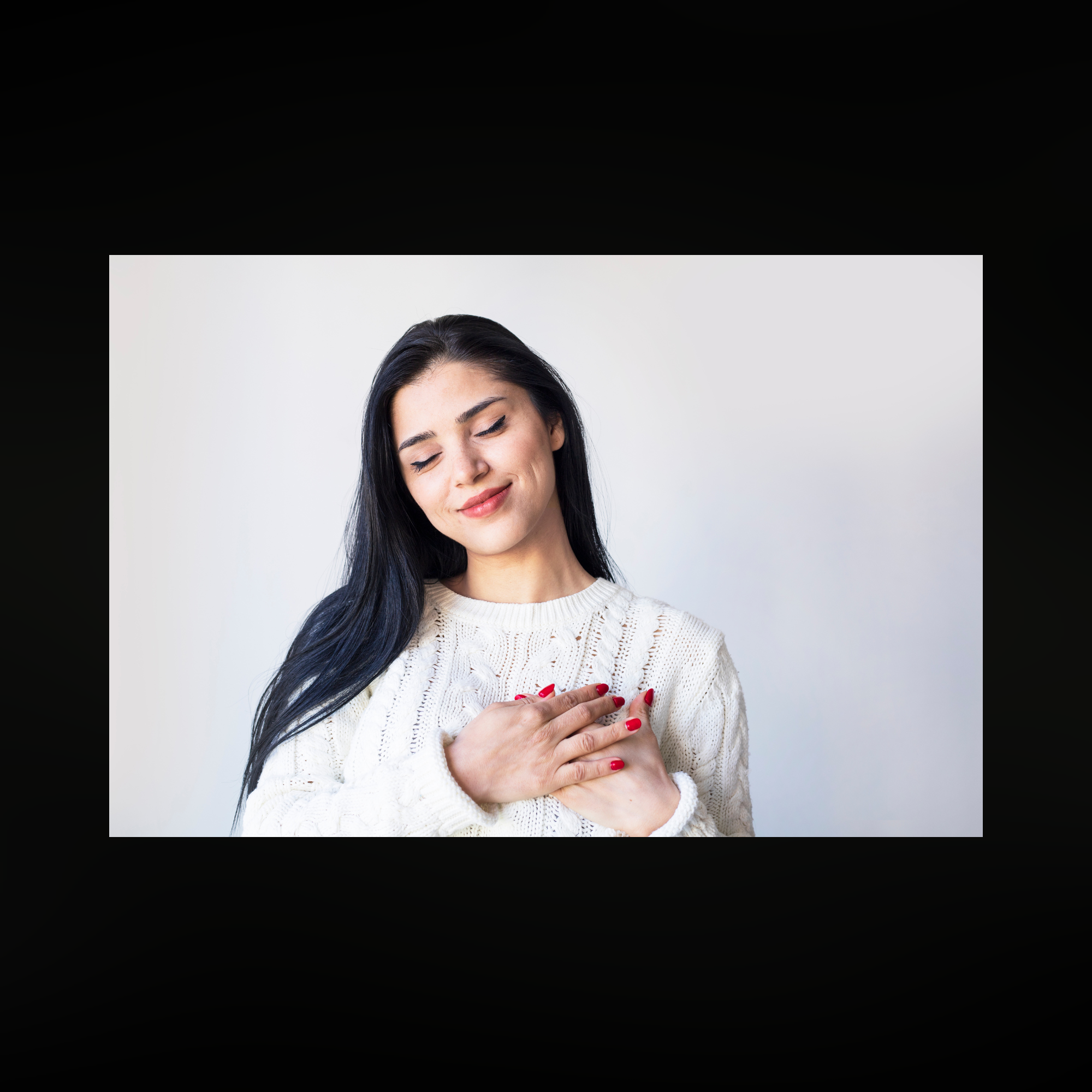 Young woman with long dark hair wearing a white cable-knit sweater, eyes closed and hands crossed over her heart, smiling gently in a minimalist setting, conveying gratitude, mindfulness, and serenity.